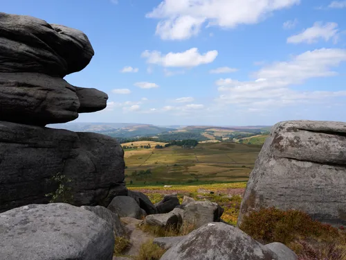This landscape photograph showcases Hope Valley in England’s Peak District, taken from between the distinctive rocks on the Longshaw Estate. The image captures rolling fields and hills typical of the United Kingdom countryside, with the expansive Hope Valley stretching into the distance. It is midday in late summer, as indicated by the bright sunlight, clear blue sky, and the lush greenery interspersed with patches of blooming heather. The rocky outcrops in the foreground frame the natural beauty of the Peak District, providing a sense of the rugged terrain for which Longshaw Estate is known. The overall scene offers a quintessential view of rural England, highlighting the iconic landscape and geology of the region.